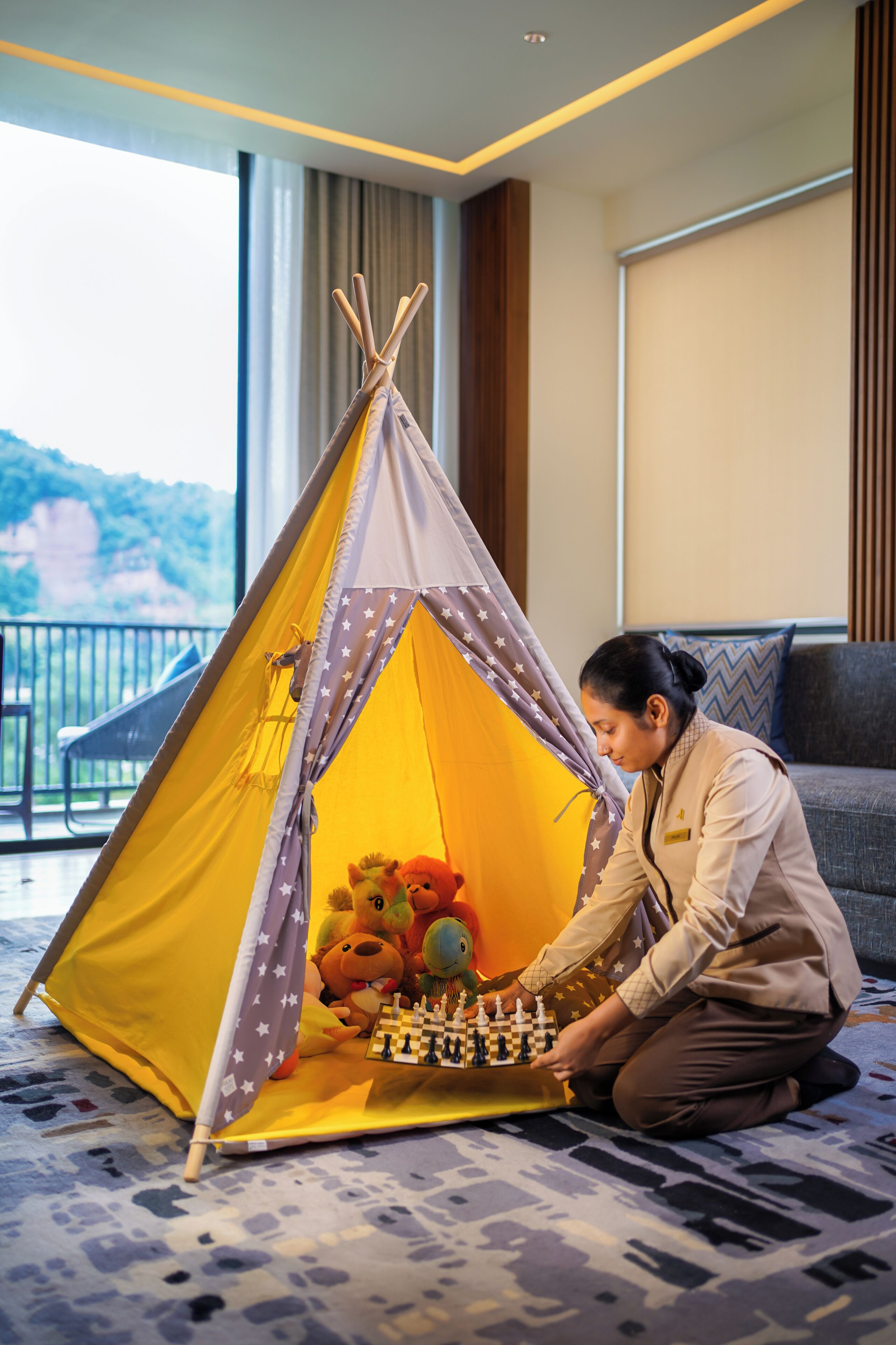 A person playing chess inside a yellow tent with star patterns, located in a room with a view of greenery.