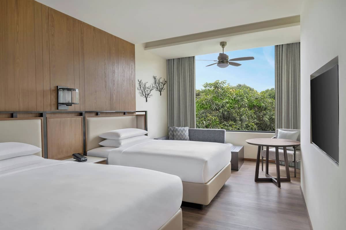 A hotel room with two beds, a small table, a sofa, a ceiling fan, and a large window with a view of greenery.
