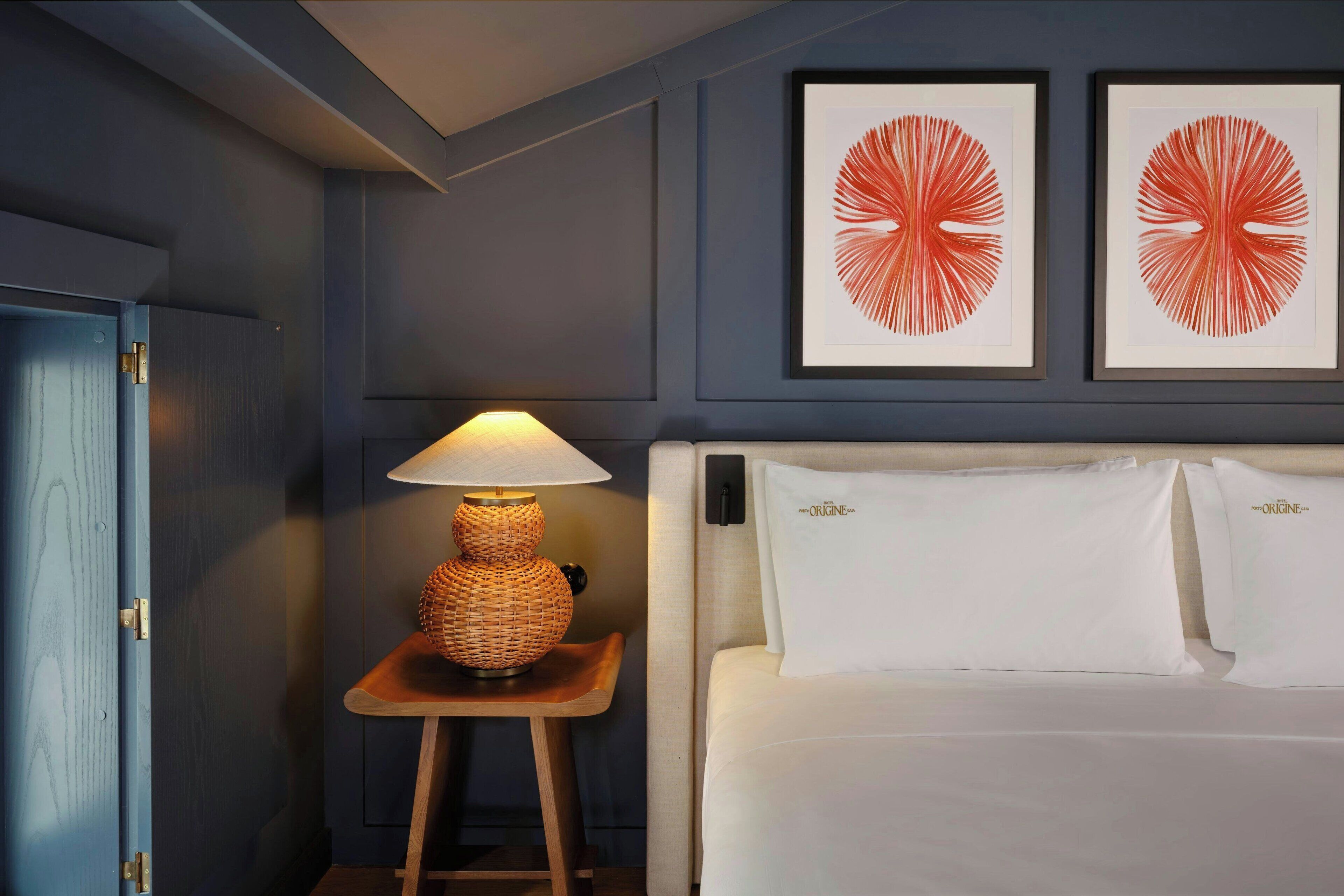 A hotel room with a bed, a nightstand with a lamp, and two framed artworks on the wall.
