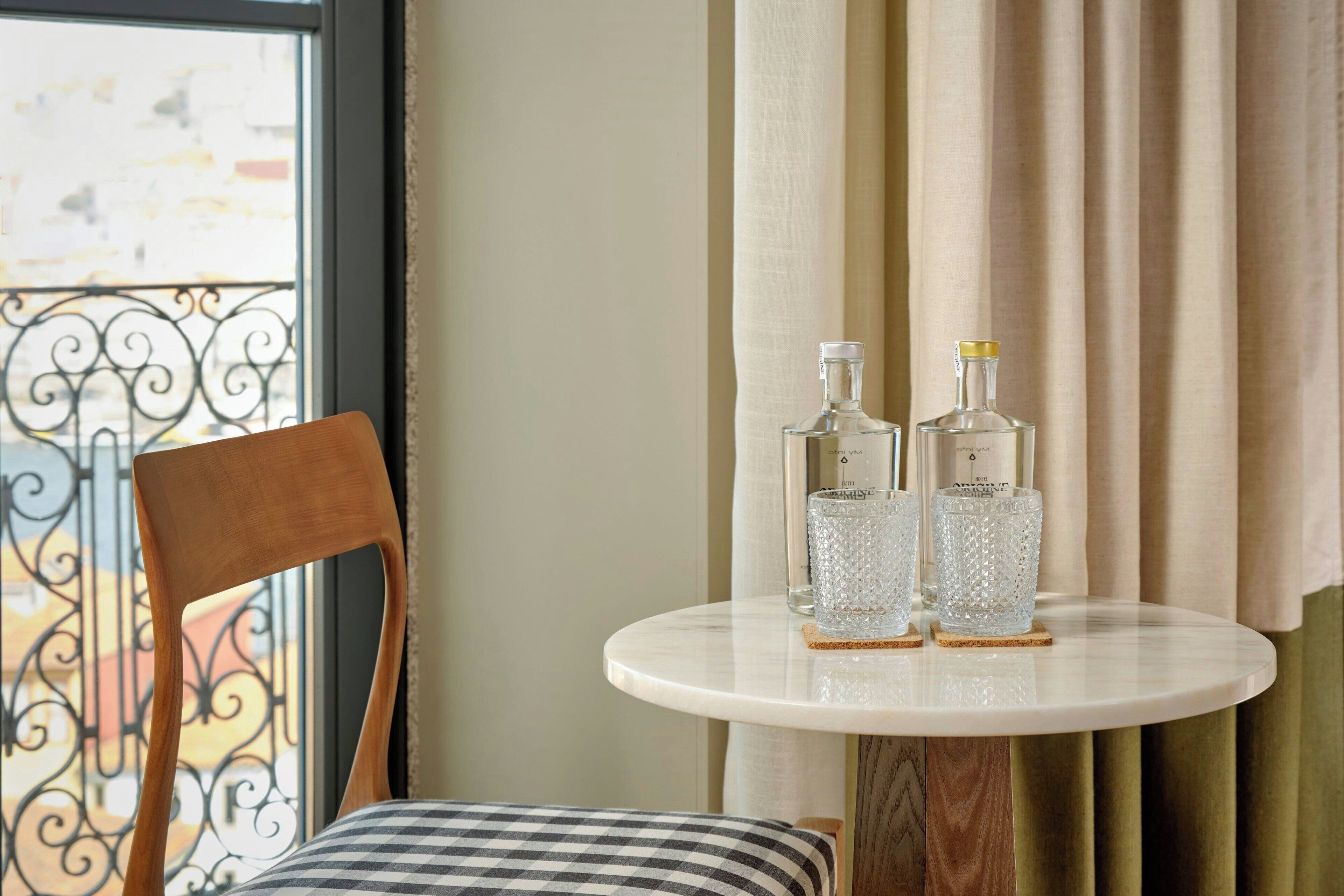 Una mesa redonda con dos botellas y dos vasos sobre posavasos, una silla de madera con cojín a cuadros, y vistas a un balcón con baranda de hierro forjado.