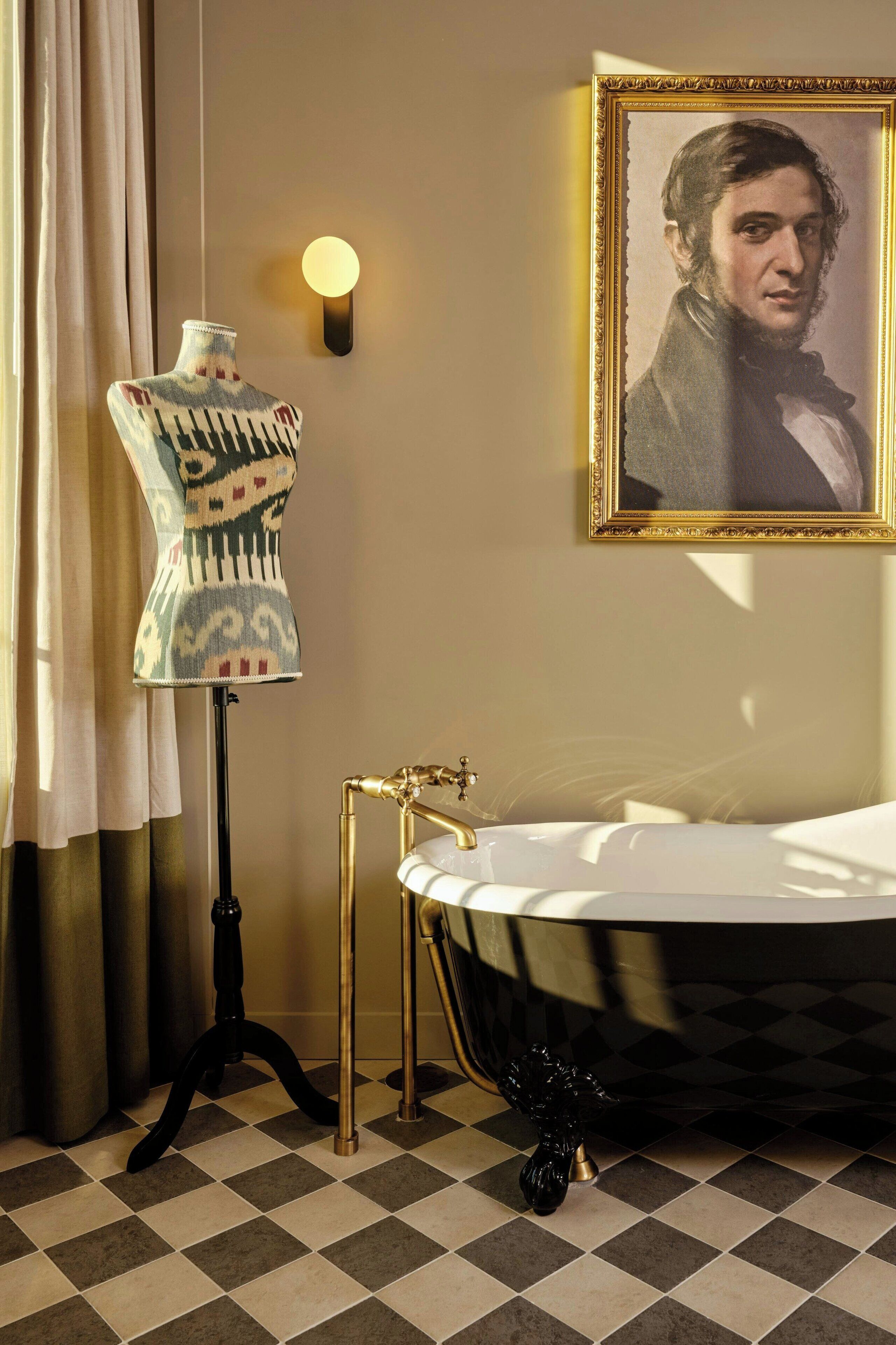 A bathroom with a checkered floor, a black bathtub, a gold faucet, a mannequin in a patterned dress, a wall-mounted light, and a framed portrait.