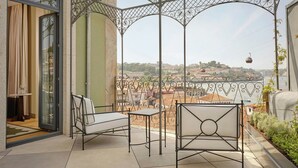 Varanda com duas cadeiras e uma mesa, pérgola de ferro forjado e vista para um horizonte urbano com edifícios e um teleférico.