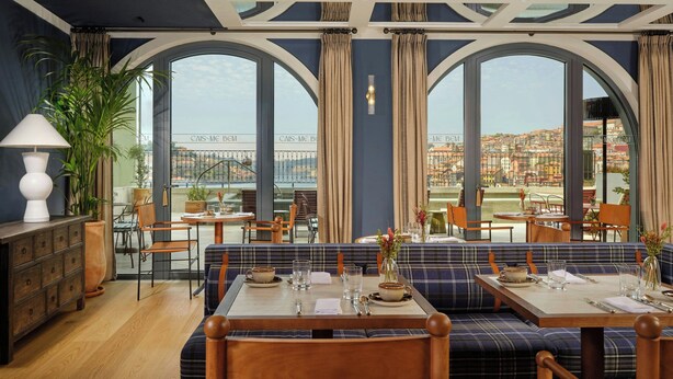 Área de jantar com vista para uma ponte e paisagem urbana, com mesa posta, pratos, copos e um vaso de flores.