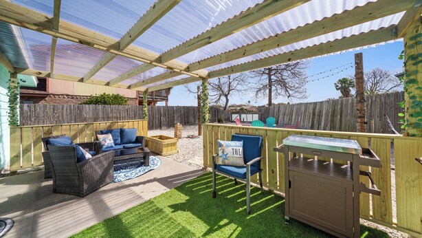 Ein überdachter Patio-Bereich mit Holzterrasse, blauem Sessel, Rattan-Sofa und Grill.