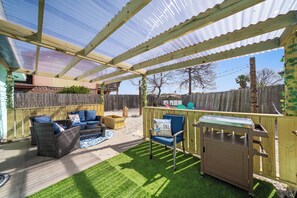 A covered patio area with a wooden deck, a blue chair, a wicker sofa, and a grill.