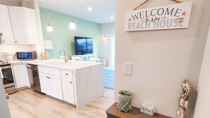 Una cocina moderna con armarios blancos, una isla central y un televisor. Se observa un letrero que dice 'Welcome to Our Beach House'.