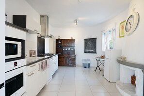Una cocina moderna con armarios blancos, un microondas, estufa y un lavaplatos. Se observa un mueble de madera con vajilla y un pequeño bote de basura.