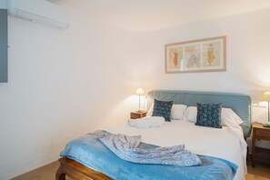Una cama bien hecha con una manta azul y almohadas estampadas, una mesita de noche con una lámpara y una obra de arte enmarcada en la pared.