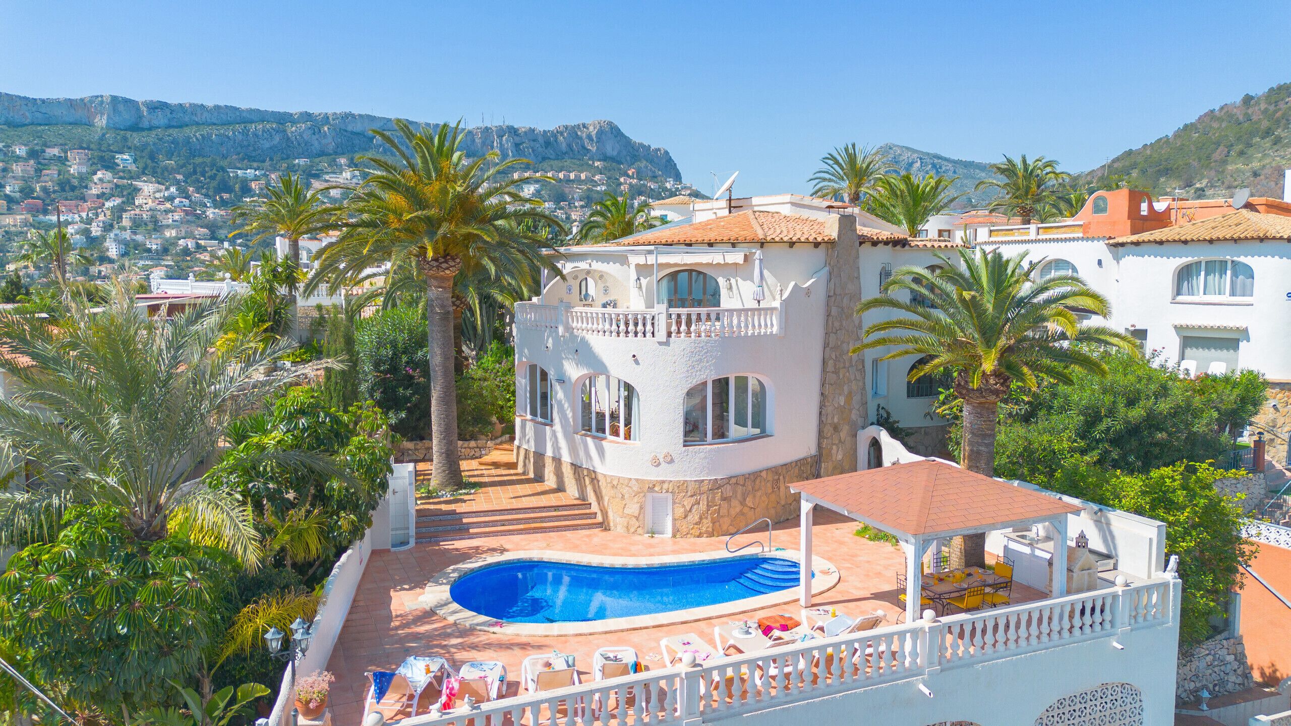 A villa with a swimming pool, surrounded by palm trees and a view of a coastal town.