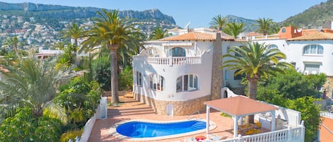 A villa with a swimming pool, surrounded by palm trees and a view of a coastal town.