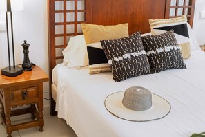 A neatly made bed with patterned pillows, a wooden nightstand with a lamp, and a woven hat on the bed.