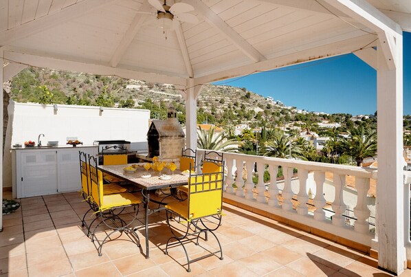 Un patio techado al aire libre con una mesa y sillas amarillas, una parrilla y vistas a un pueblo en la ladera.