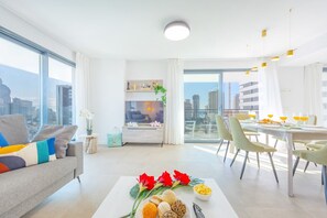 A modern living room with a sofa, dining table, and chairs, a TV, and a balcony with a city view.