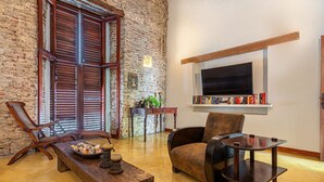 A cozy room with a brick wall, wooden shutters, a bookshelf, a television, and a dining area with a table and chairs. - Casa Palos - Old City - Luxury (Cartagena)