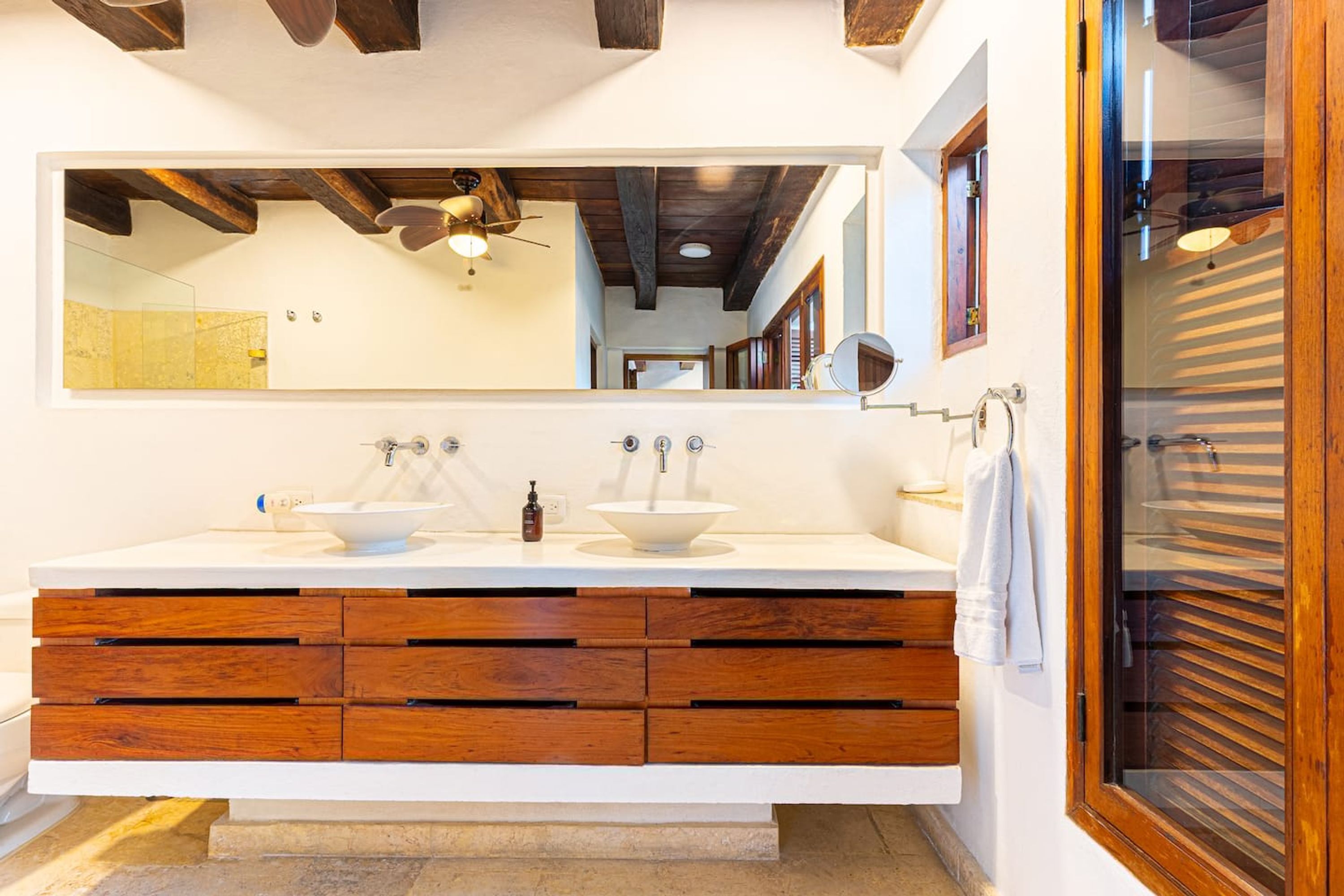 A bathroom with a large mirror, two sinks, wooden drawers, and a ceiling fan.