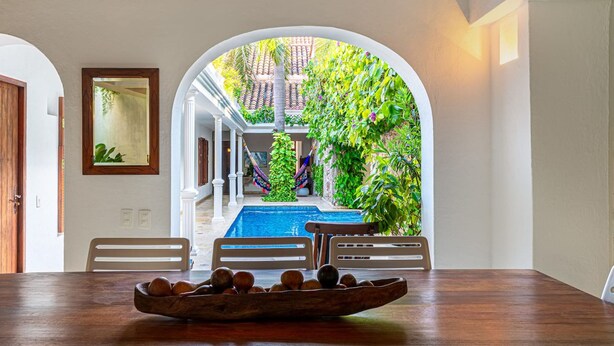 A dining area with a wooden table and chairs, a bowl of fruit, and a view of a pool and outdoor area with plants and a hammock. - Casa Palos - Old City - Luxury (Cartagena)