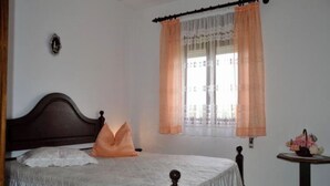 Ein Schlafzimmer mit Bett, einem Fenster mit Vorhängen, einem kleinen Tisch mit Blumen und einer Wandleuchte.