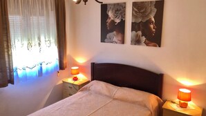 Een slaapkamer met een bed, twee nachtkastjes met lampen, een raam met gordijnen en twee bloemschilderijen aan de muur.