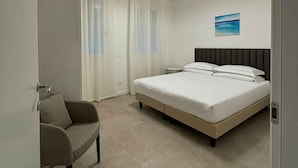 Una habitación de hotel con una cama, una silla, un cuadro y un ventanal con cortinas.