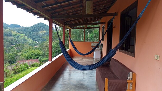 A balcony with a hammock, a sofa, and a view of green hills. - Little house in the mountains (Gonçalves)