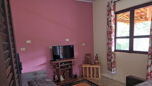 A living room with a television on a wooden stand, a small table with decorative items, and a window with floral curtains. - Little house in the mountains (Gonçalves)