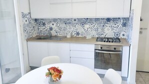 A compact kitchen with white cabinets, a blue and white patterned backsplash, a gas stove, and a small dining area with a white table and chairs. - CN001 - Cesenatico, two-room apartment with balcony and air conditioning (Cesenatico)