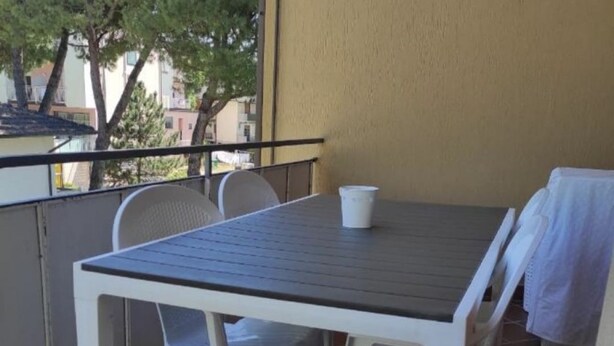 A balcony with a table and chairs, a white bucket, and a view of buildings and trees. - CN001 - Cesenatico, two-room apartment with balcony and air conditioning (Cesenatico)