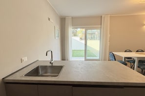 A kitchen with a sink, a dining table, chairs, and a view of a green lawn and building through a glass door.