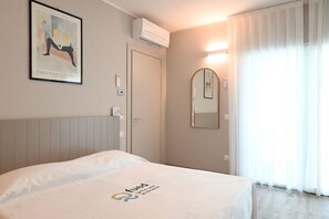 A hotel room with a bed, a wall-mounted air conditioner, a door, a framed picture, a mirror, and a window with curtains.