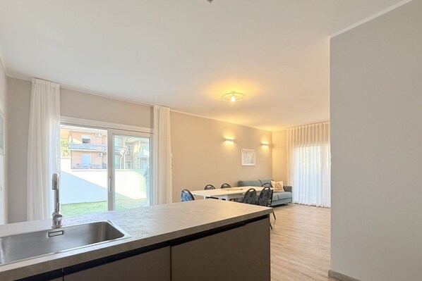A modern kitchen with a sink, dining table, and chairs, a balcony with a view, and a living area with a sofa and a TV.