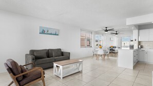 A modern living room with a grey sofa, a wooden chair, and a coffee table. There is a kitchen area with white cabinets and a dining table with chairs. - Bay Harbor Sunset Apt 4A (Bay Harbor Islands)