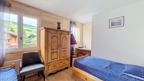 Un dormitorio con un armario de madera, una cama individual con cubrecama de lunares, una silla y una ventana con vistas a casas y follaje.