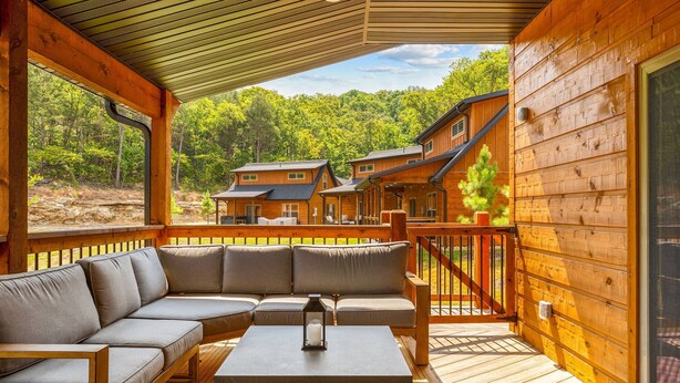 A wooden deck with a sofa and table, overlooking a forested area with cabins.