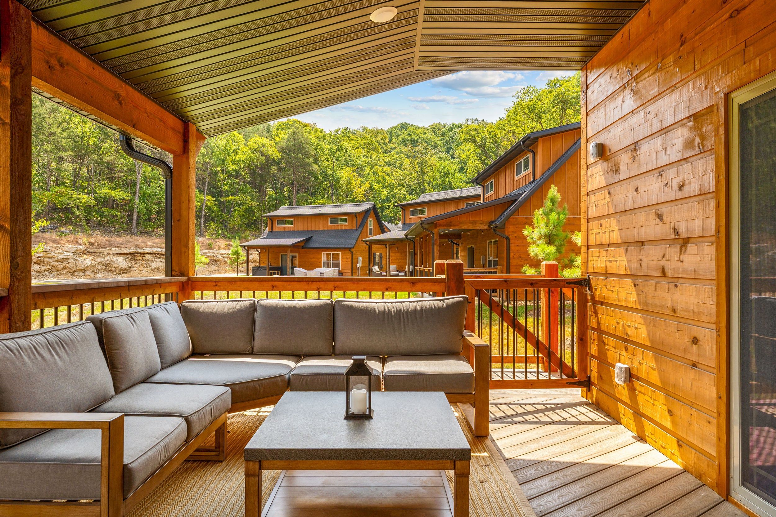 A covered outdoor seating area with a sofa and table, overlooking a forested landscape and a row of cabins.