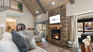 A spacious living room with a stone fireplace, a high ceiling with exposed beams, and a balcony with a railing.