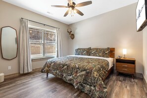Ein Schlafzimmer mit einem Bett, einem Deckenventilator, einem aufgehängten Hirschgeweih, einem Fenster mit Vorhängen und einem Nachttisch mit Lampe.