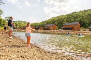 Une scène au bord d’un lac, avec des cabanes en bois, une personne pratiquant le kayak et une autre personne qui pêche.