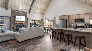 A spacious living area with a stone fireplace, a large sofa, and a dining table with chairs. A kitchen area with modern appliances and bar stools is visible.