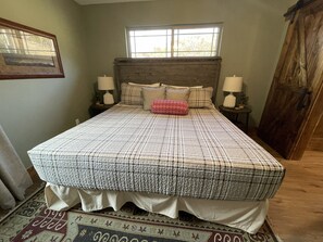 Un dormitorio con una cama grande, dos mesitas de noche con lámparas, una ventana sobre la cama y una puerta de madera.