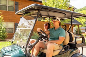 A golf cart with two people inside, parked in front of a wooden house with a porch.