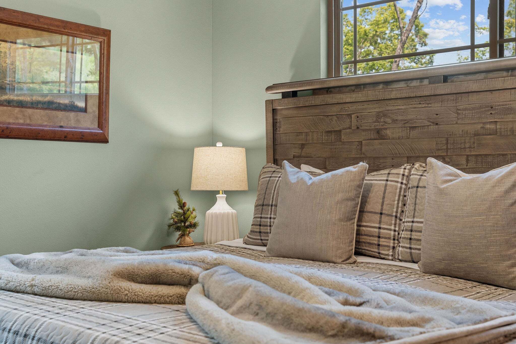 A bedroom with a wooden headboard, a bed with multiple pillows, a bedside lamp, and a framed picture on the wall.