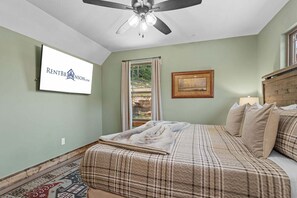 Un dormitorio con cama, ventilador de techo, ventana con cortinas, un cuadro enmarcado y un cartel en la pared.