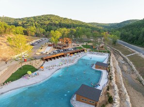 A large outdoor swimming pool with a sandy beach area, surrounded by lounge chairs and umbrellas. There are several buildings with wooden structures and a parking area with cars.
