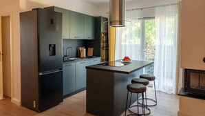 Eine moderne Küche mit dunklen Schränken, einem Edelstahl-Kühlschrank, einer Kochinsel mit Sitzgelegenheiten und einem großen Fenster mit durchsichtigen Vorhängen.