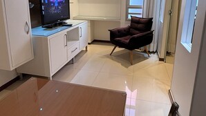 Una habitación de hotel moderna con una mesa de comedor de madera, un televisor de pantalla plana, un sillón cómodo y un amplio ventanal con cortinas.