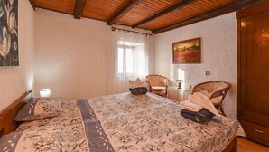 Une chambre avec un plafond en bois, un lit recouvert d’une literie à motifs floraux, deux fauteuils en osier, une petite table avec une lampe et un tableau accroché au mur.