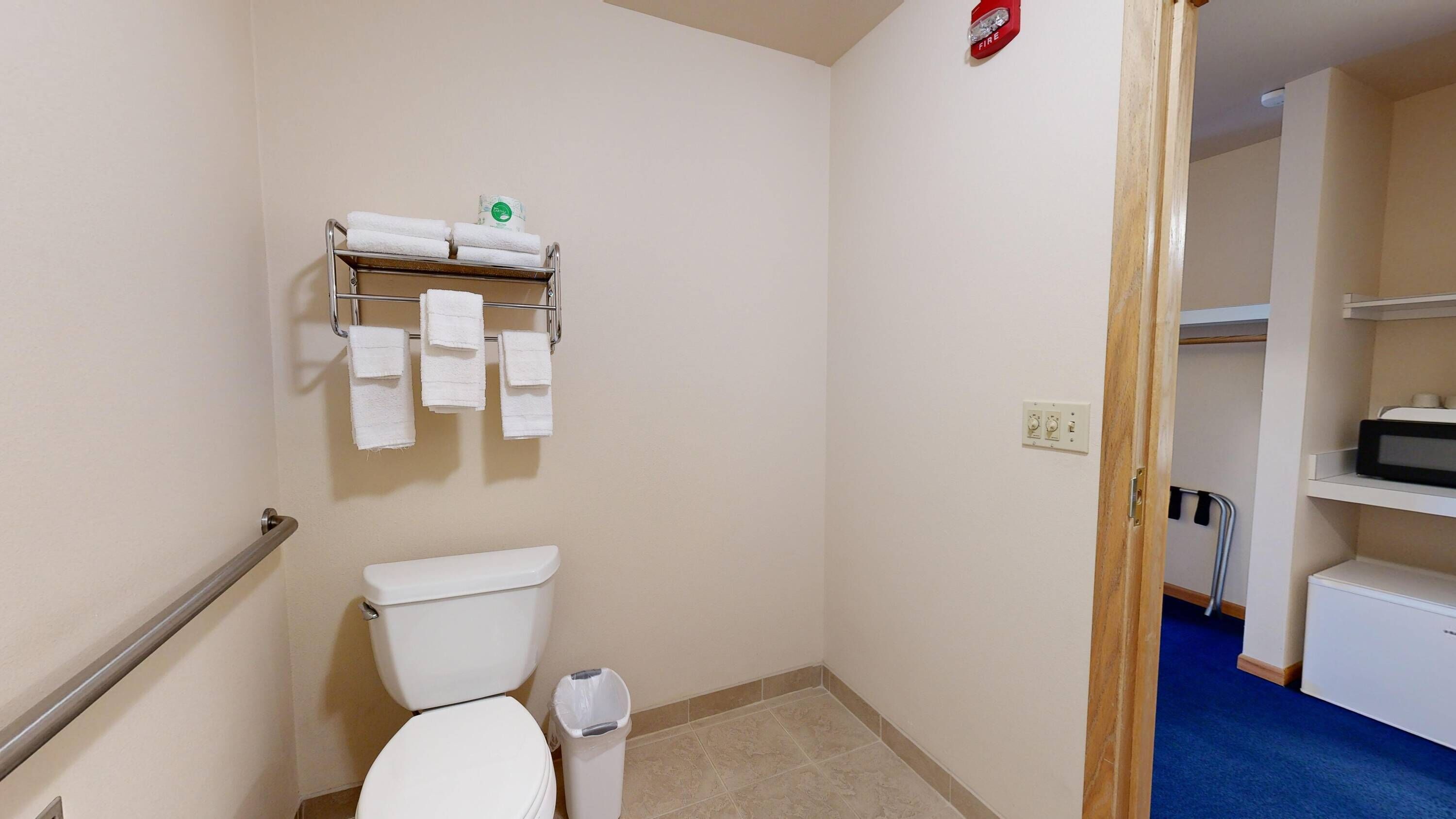 A small bathroom with a toilet, a towel rack with towels, a trash bin, and a fire alarm.