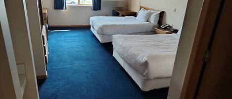 A hotel room with two beds, a blue carpet, a window with curtains, an air conditioning unit, and a bedside table with a lamp.