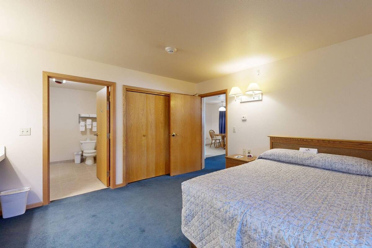 Ein Schlafzimmer mit Bett, Holz-Kopfteil, Nachttisch, Schreibtisch und einem durchgehenden Türspalt sichtbaren Badezimmer.