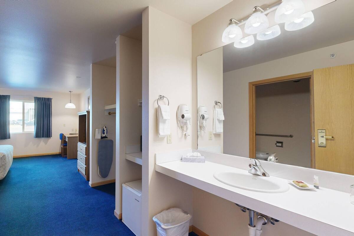 Ein Hotelzimmer mit Bett, Schreibtisch, Kleiderschrank, einem Badezimmer mit Waschbecken und Spiegel sowie einem blauen Teppich im Flur.